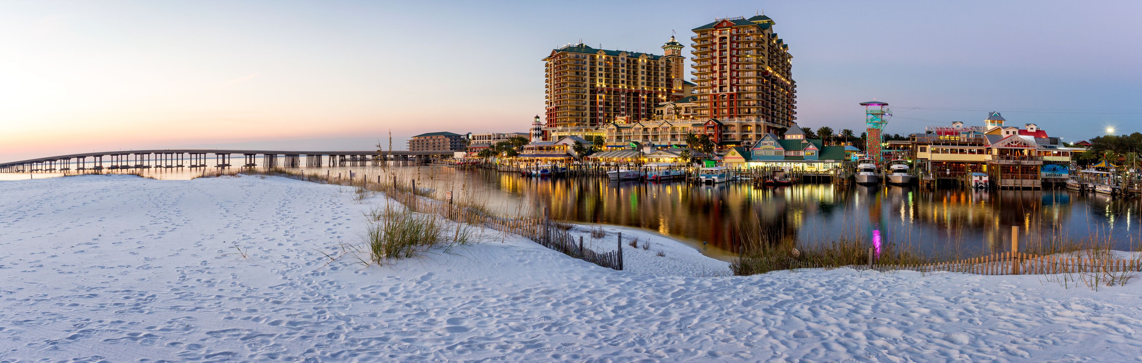 A snowy beach with a pier, boats, and buildings reflecting on the water.
