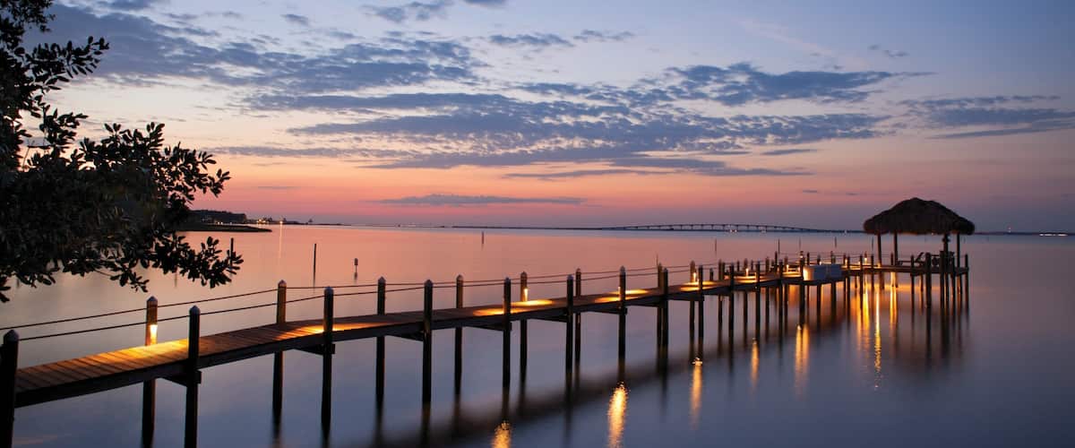 A wooden pier extending into calm waters at sunset, with a thatched-roof structure at the end.