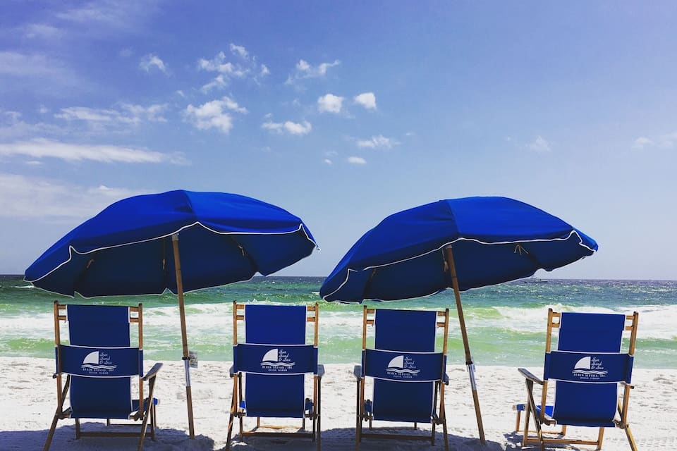 Four blue beach chairs and umbrellas on a sandy beach with the ocean in the background.