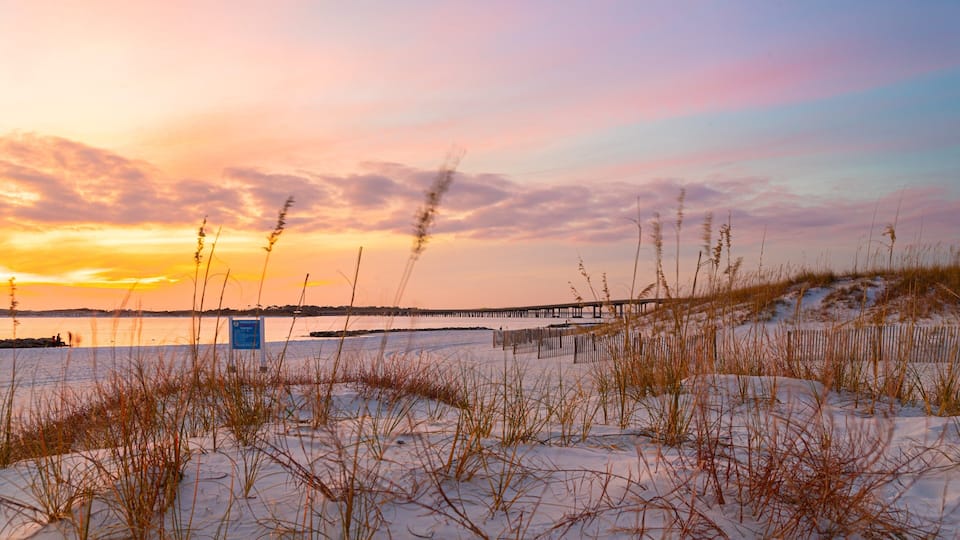 A sunset over a beach with a wooden pier and sand dunes.