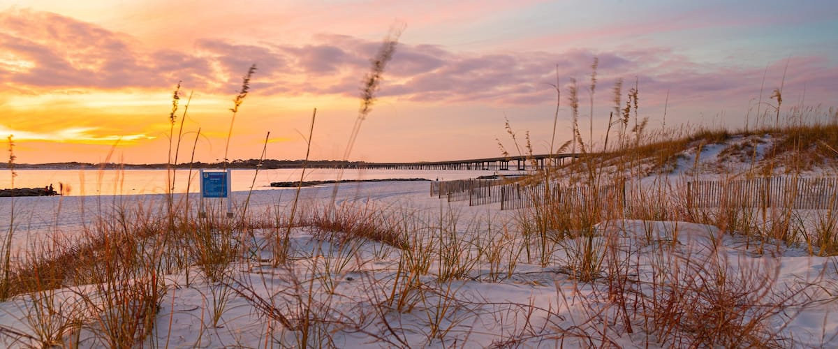 A sunset over a beach with a wooden pier and sand dunes.
