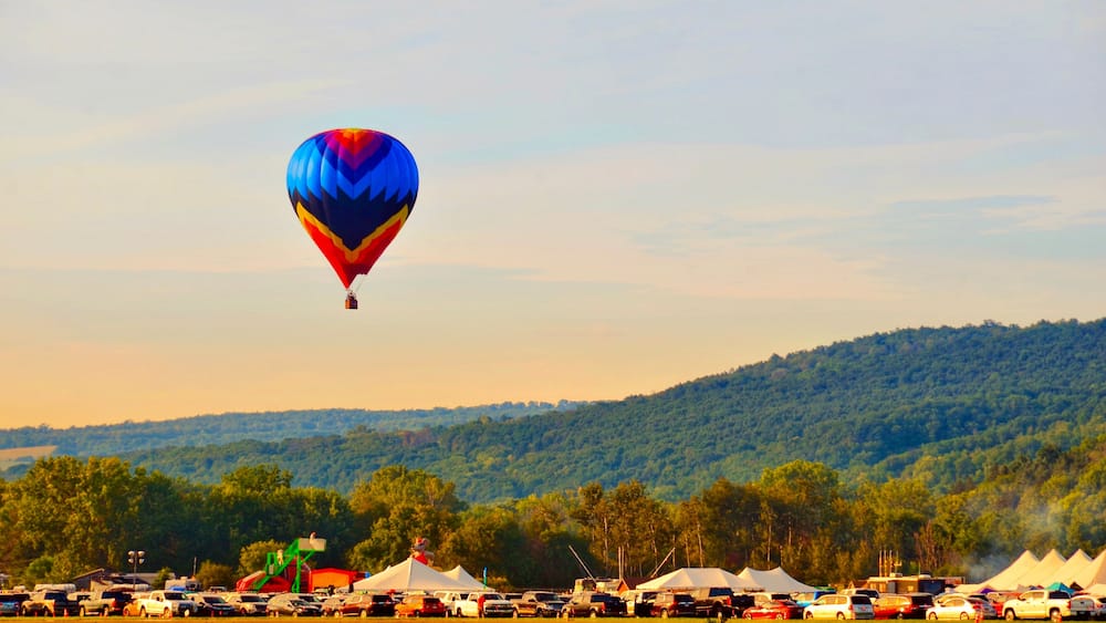 A colorful hot air balloon flying over a parking lot with tents and vehicles, set against a backdrop of a forested hill.