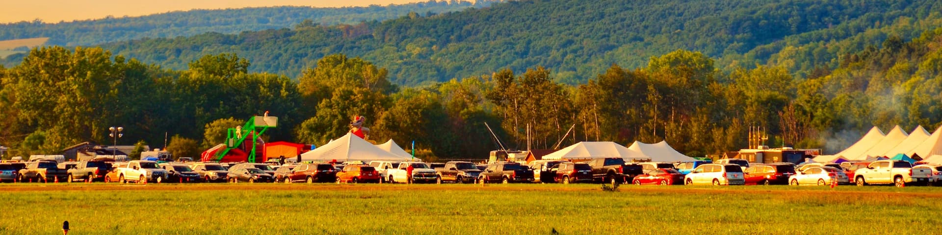 A colorful hot air balloon flying over a parking lot with tents and vehicles, set against a backdrop of a forested hill.