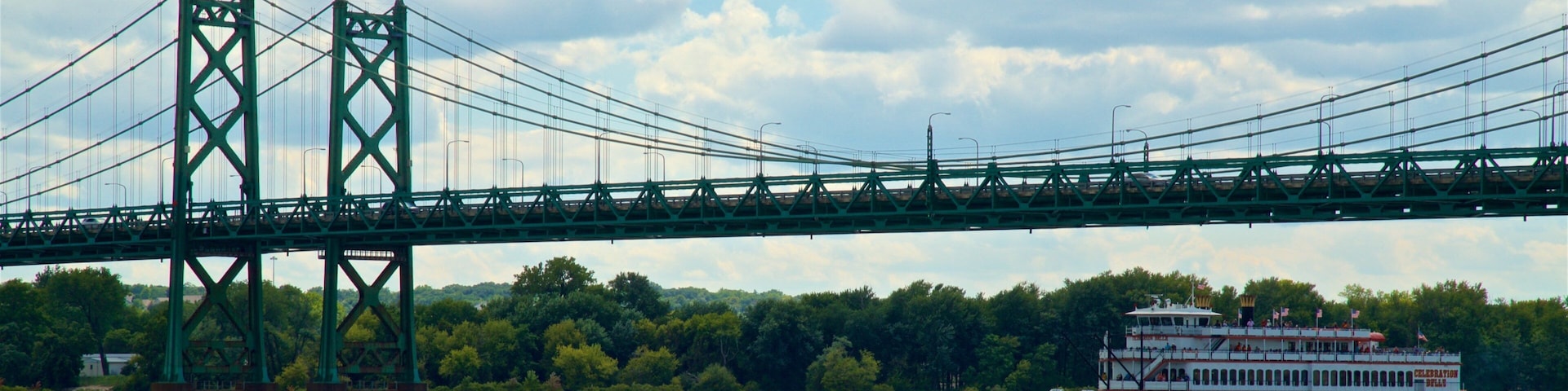 A large green bridge spanning a river with a white riverboat docked on the right.