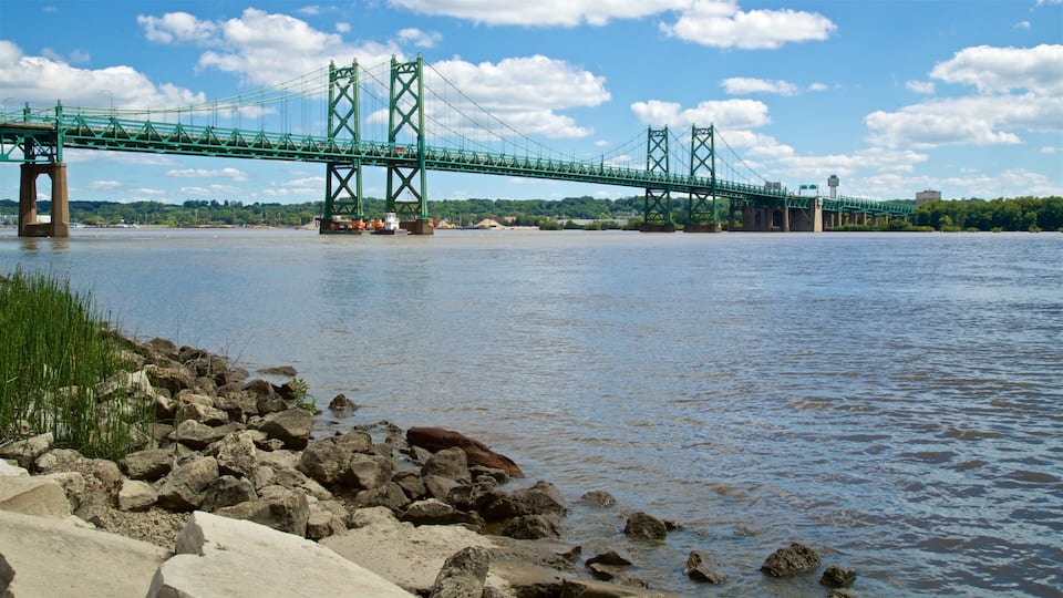 A green suspension bridge over a river with rocks and grass in the foreground.