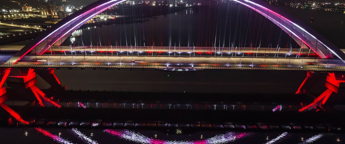 Uma ponte bem iluminada, com um arco característico e cabos iluminados, atravessa um corpo de água.