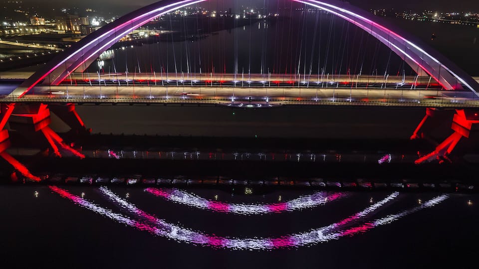 A brightly lit bridge with a distinctive arch and illuminated cables, spanning over a body of water.