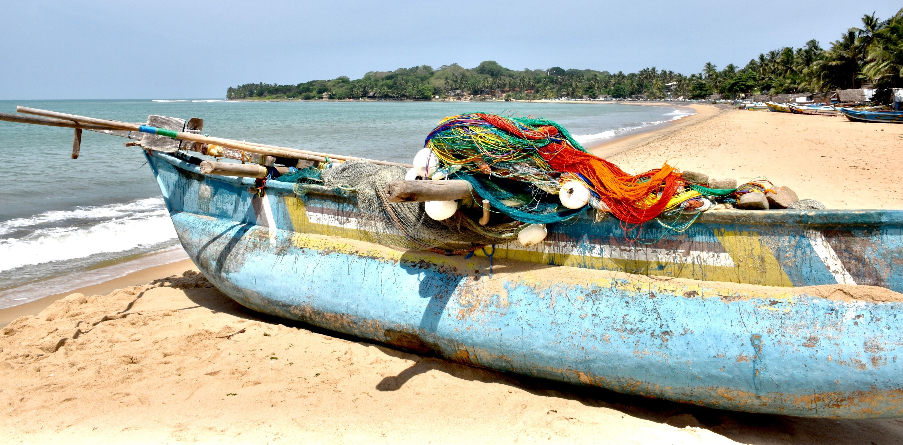 A weathered boat with colorful nets on a sandy beach, palm trees, and calm ocean in the background.