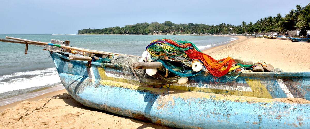 A weathered boat with colorful nets on a sandy beach, palm trees, and calm ocean in the background.