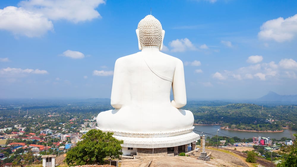 Une grande statue blanche représentant une figure assise, coiffée d’une sorte de couvre-chef arrondi, et entourée d’un socle circulaire, dominant une ville et une rivière.