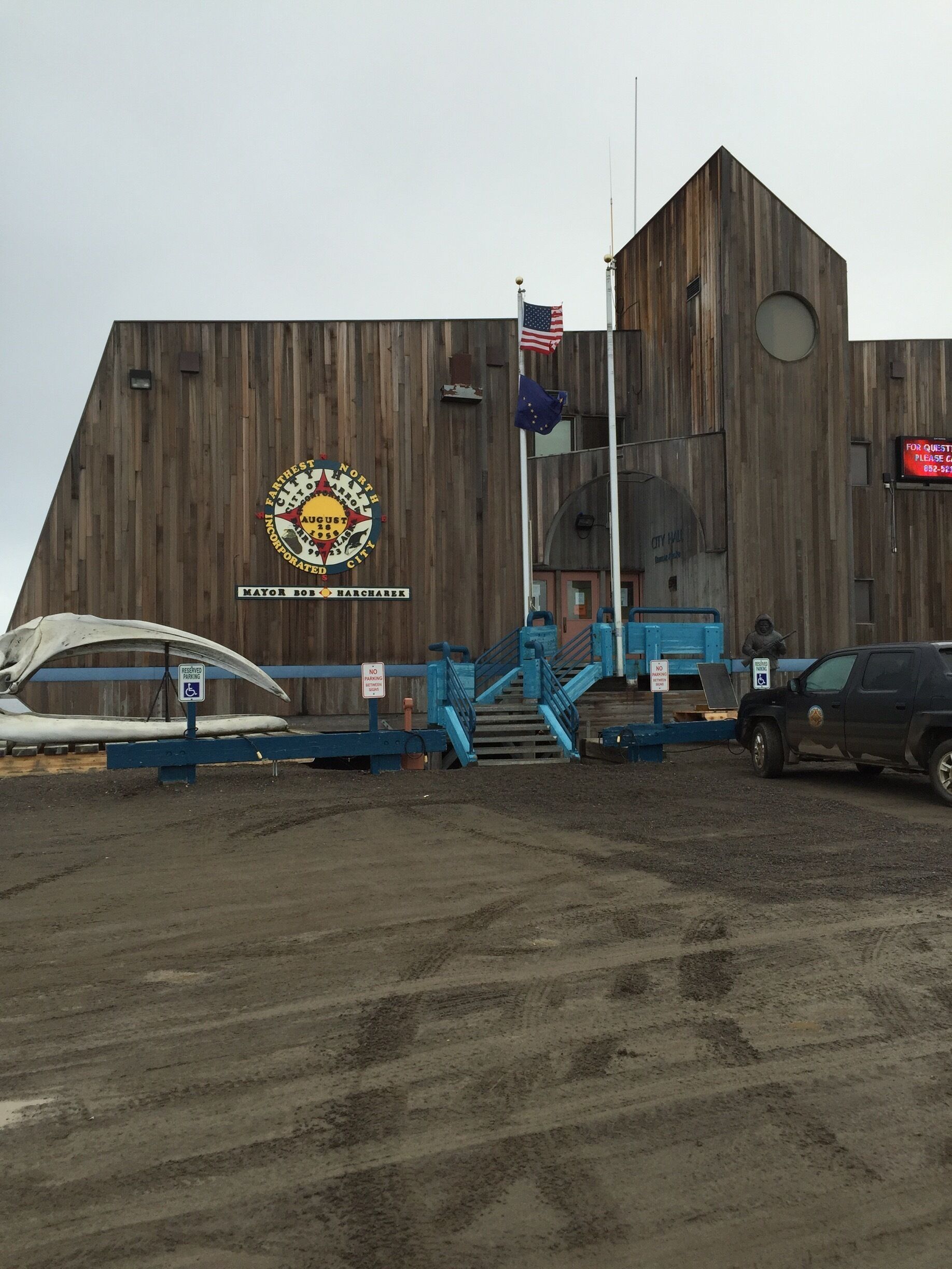 A large wooden building with a flagpole, a sign for Mayor Bob Harcharek, and a 'Reserved Parking' sign.