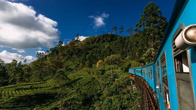 A train on a track through a lush, green landscape with a blue sky and clouds.