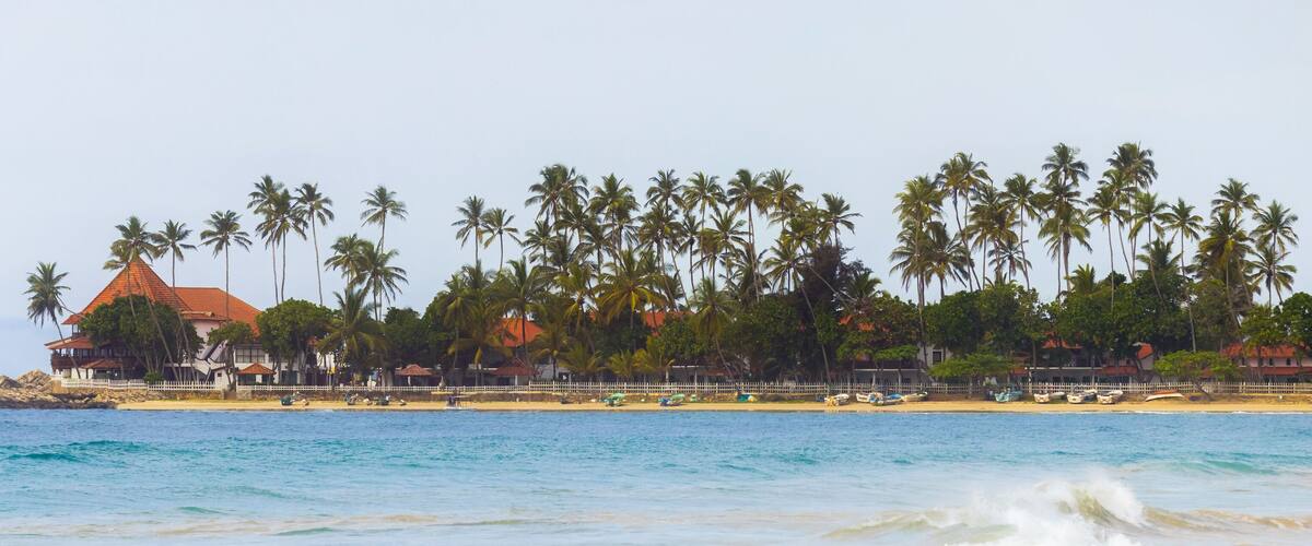 A beach with palm trees, a building with a red roof, and boats on the shore.