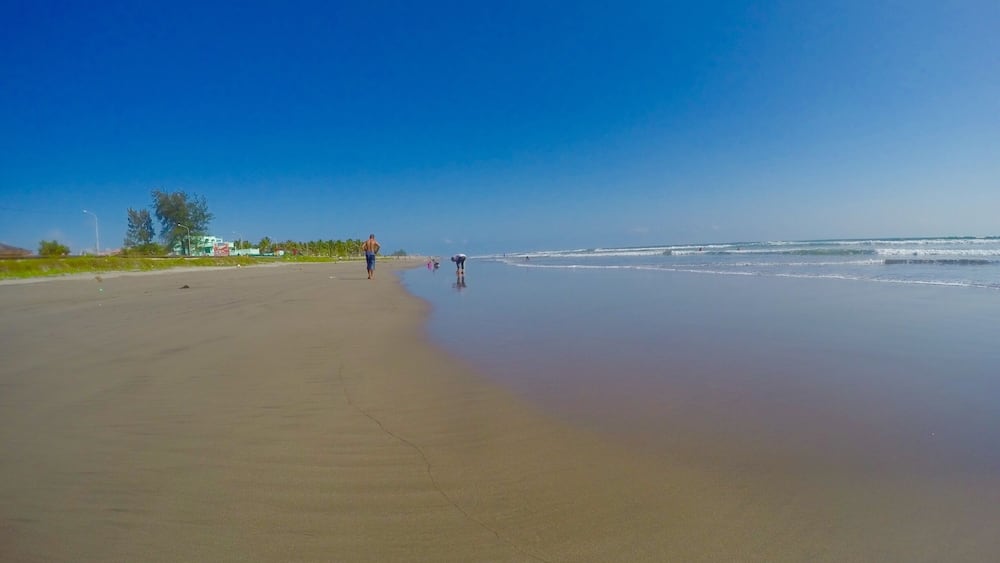 Une vaste plage de sable fin, avec de douces vagues, un ciel bleu clair et quelques personnes se promenant le long du rivage.