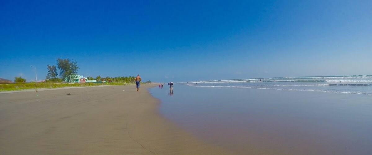 A wide sandy beach with gentle waves, a clear blue sky, and a few people walking along the shore.