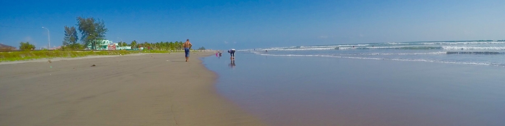 A wide sandy beach with gentle waves, a clear blue sky, and a few people walking along the shore.