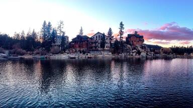 A lakeside view with a large wooden house and several smaller buildings, surrounded by trees and rocks.