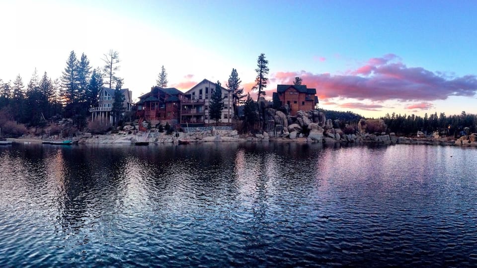 A lakeside view with a large wooden house and several smaller buildings, surrounded by trees and rocks.