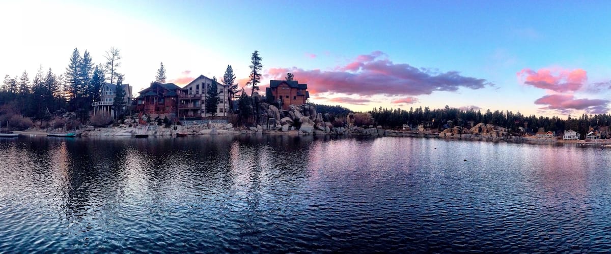 A lakeside view with a large wooden house and several smaller buildings, surrounded by trees and rocks.