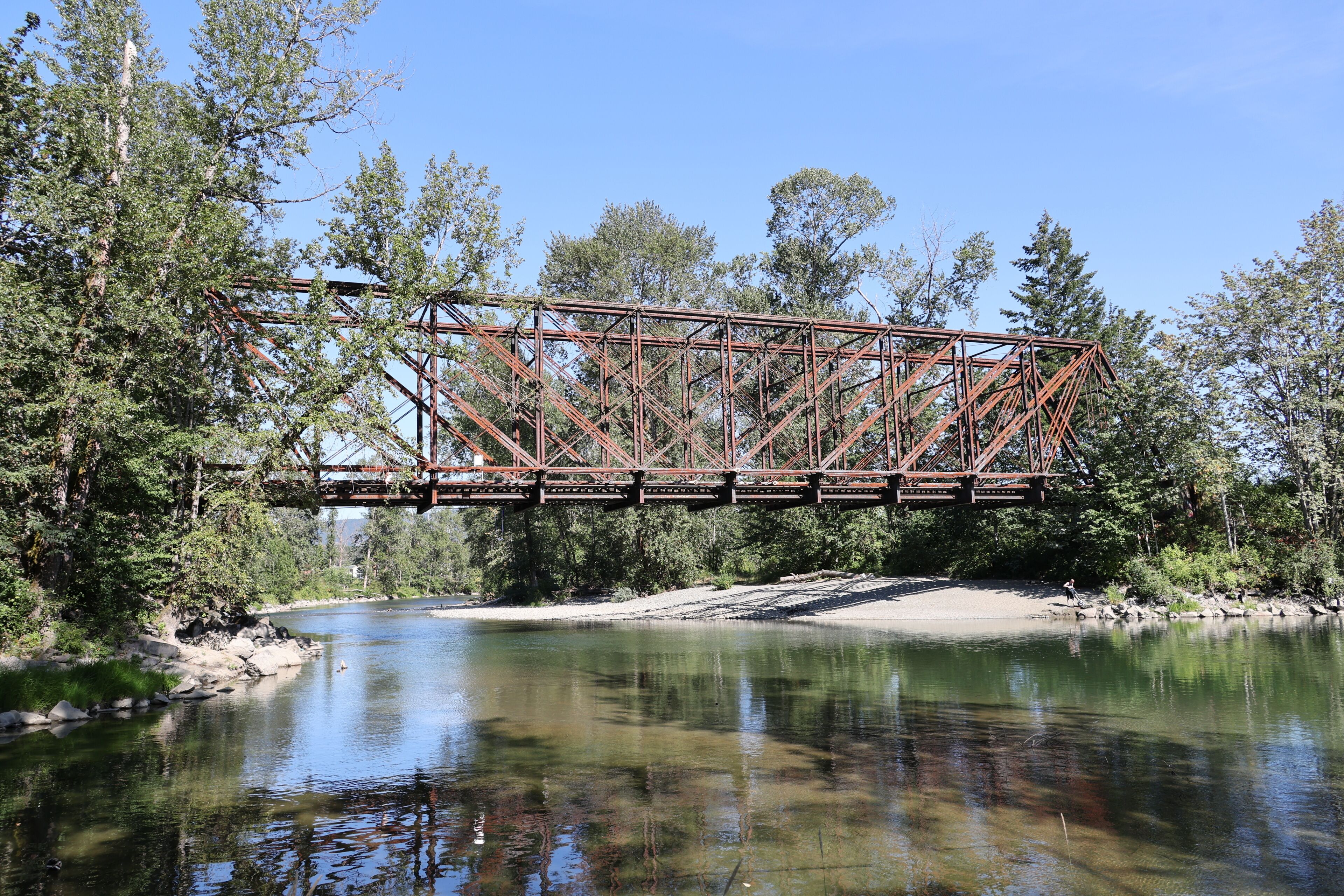 A rusted metal bridge spanning a river, surrounded by dense trees and clear water.
