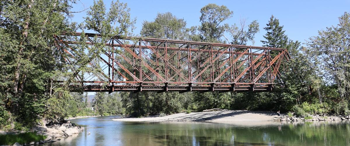 A rusted metal bridge spanning a river, surrounded by dense trees and clear water.