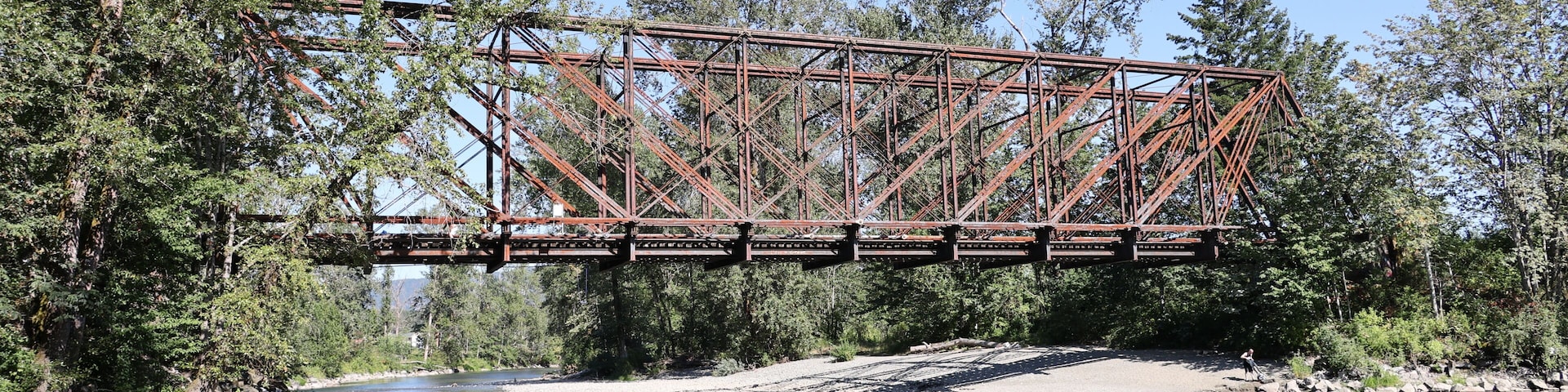 A rusted metal bridge spanning a river, surrounded by dense trees and clear water.