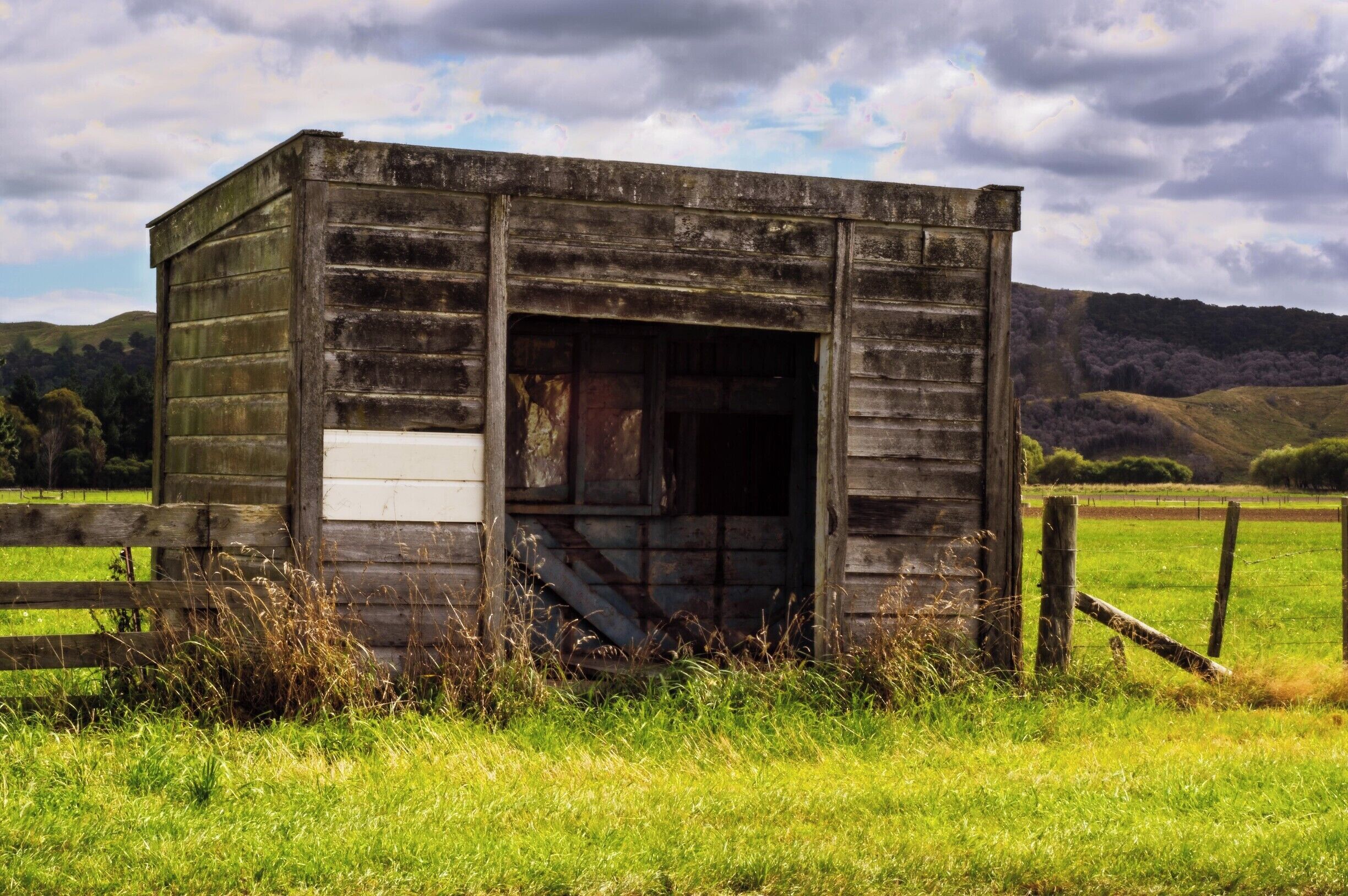 An old wooden structure with a weathered exterior, surrounded by a grassy field and a wooden fence.
