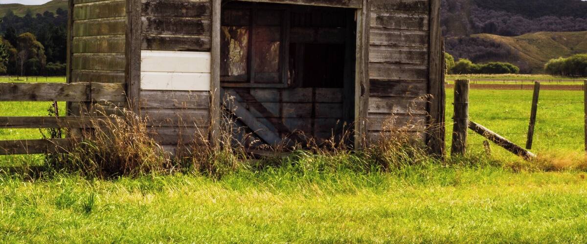 An old wooden structure with a weathered exterior, surrounded by a grassy field and a wooden fence.