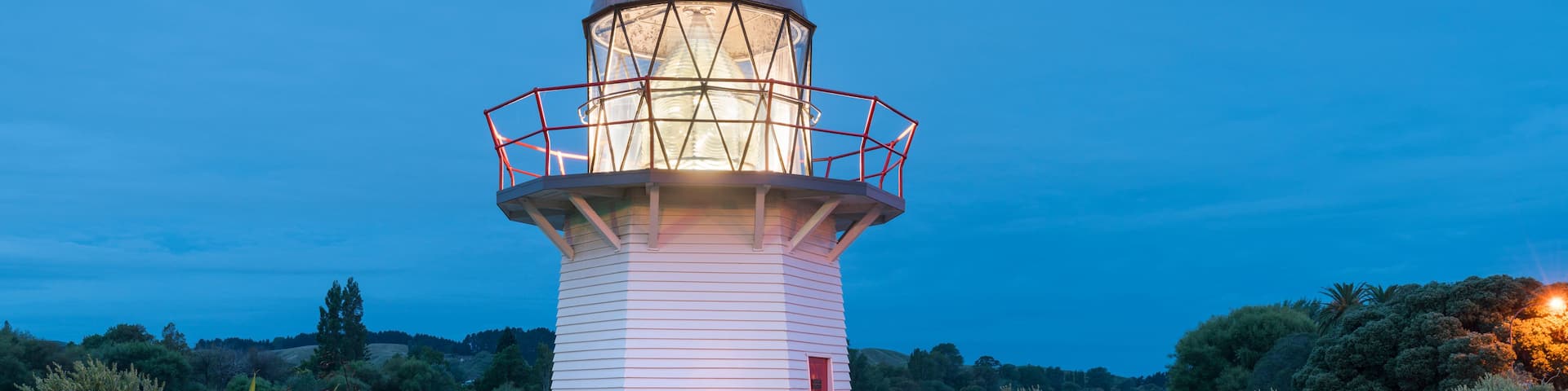 A lighthouse with a red door, surrounded by rocks and greenery, overlooking a body of water.