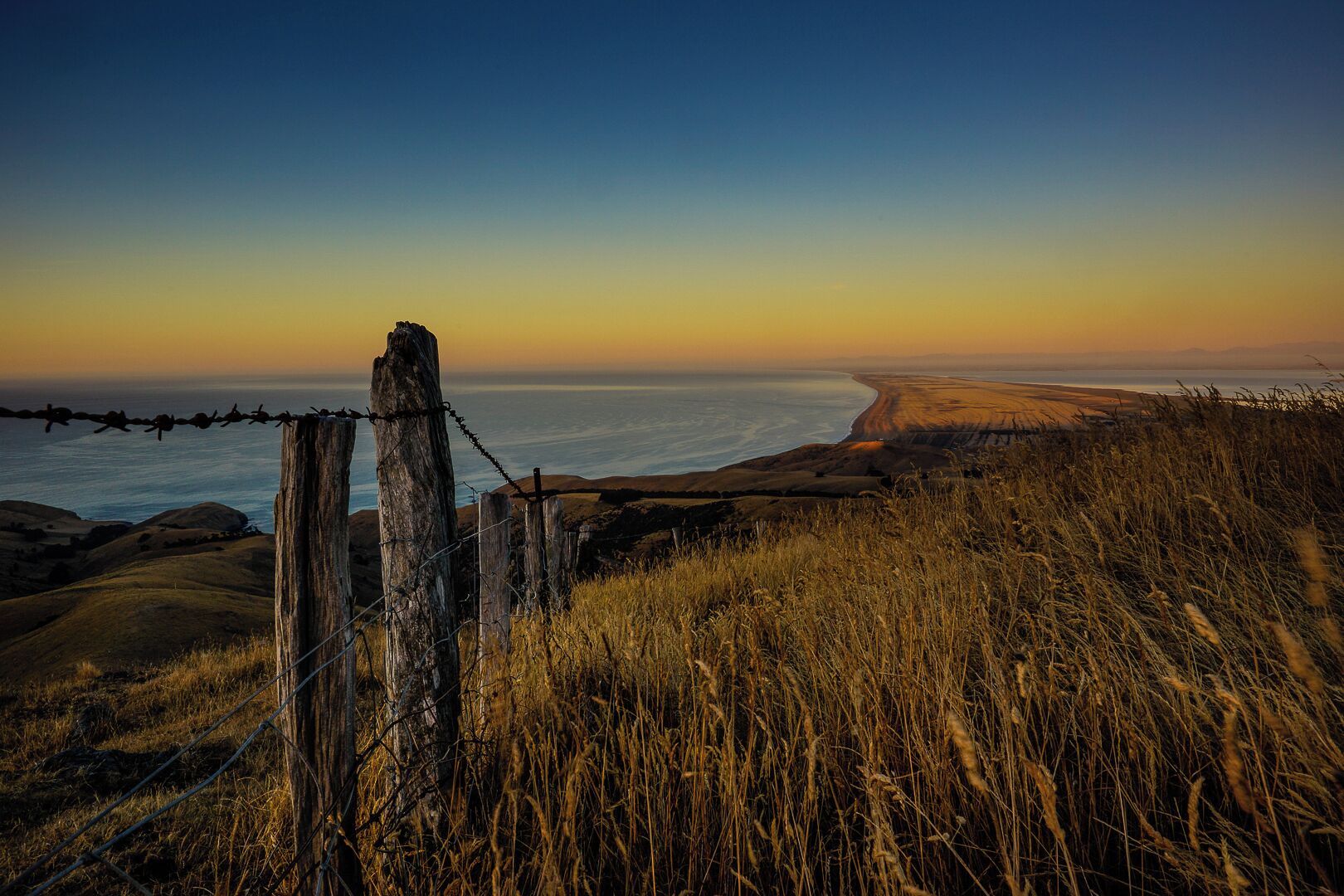 A coastal landscape with a barbed wire fence, grassy terrain, and a view of the ocean at sunset.