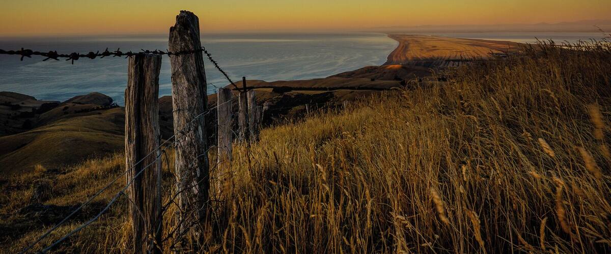 A coastal landscape with a barbed wire fence, grassy terrain, and a view of the ocean at sunset.