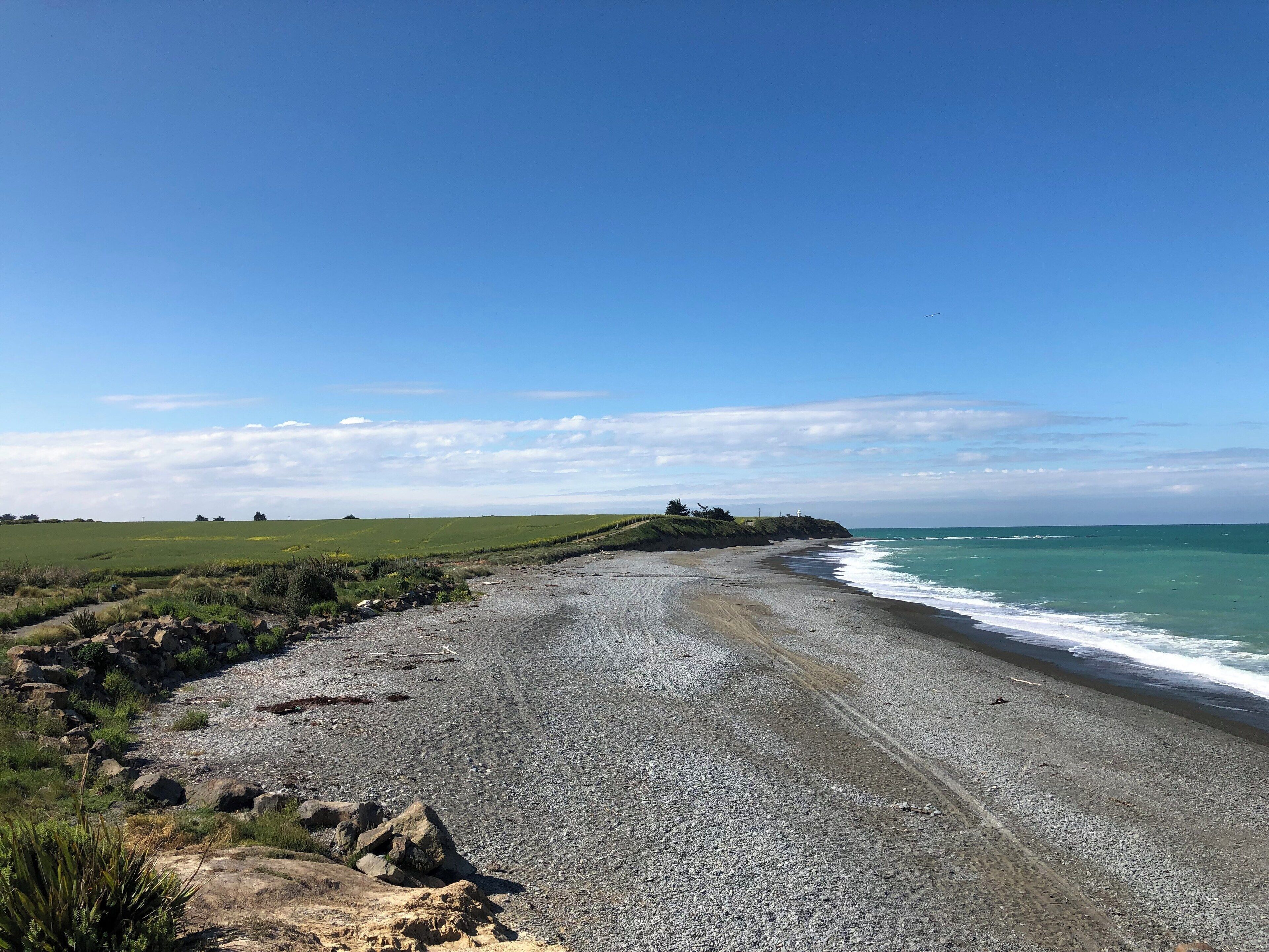 A coastal landscape with a pebbly beach, grassy fields, and a lighthouse in the distance.