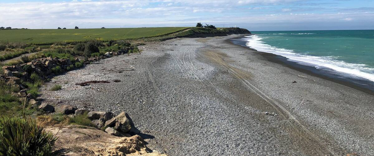 A coastal landscape with a pebbly beach, grassy fields, and a lighthouse in the distance.