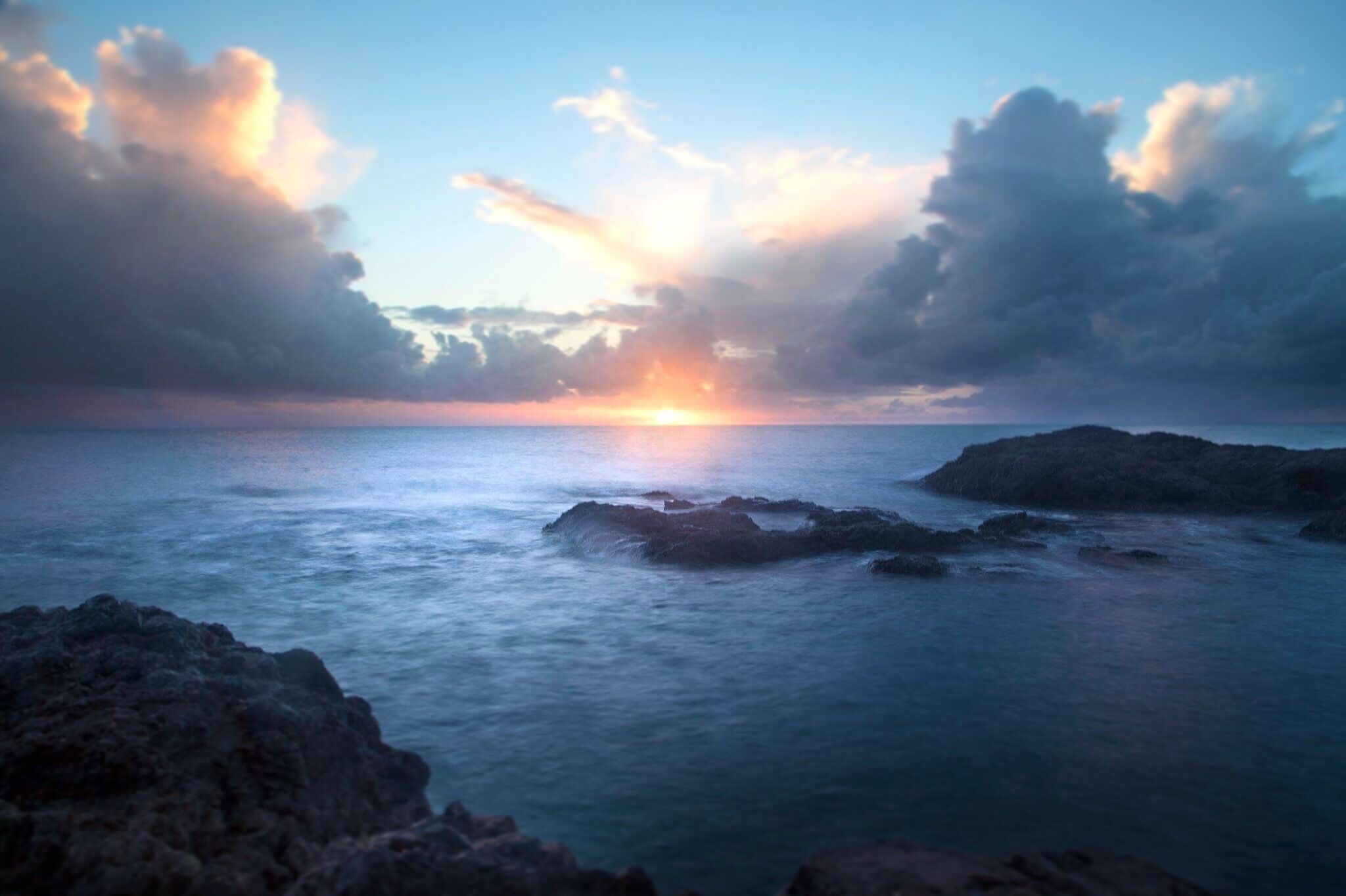 A sunset over a rocky coastline with calm sea and clouds.