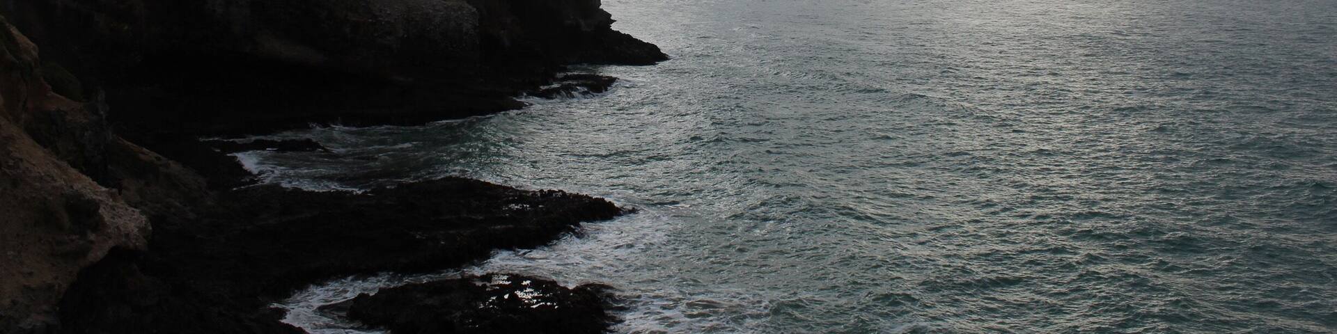 A coastal cliff with a lighthouse, rocky shoreline, and a cloudy sky.