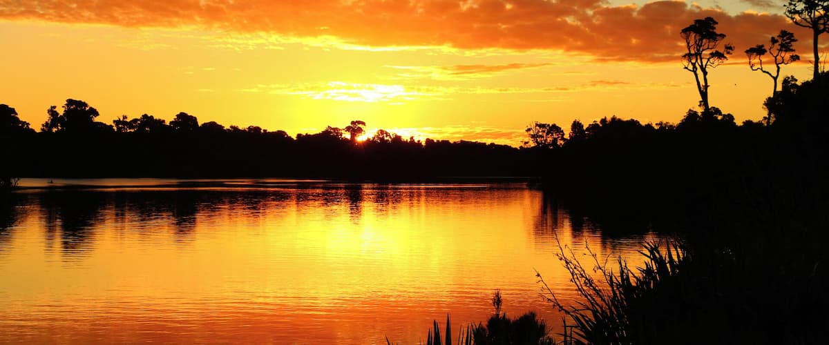 A sunset over a calm lake with silhouettes of trees and a clear sky.