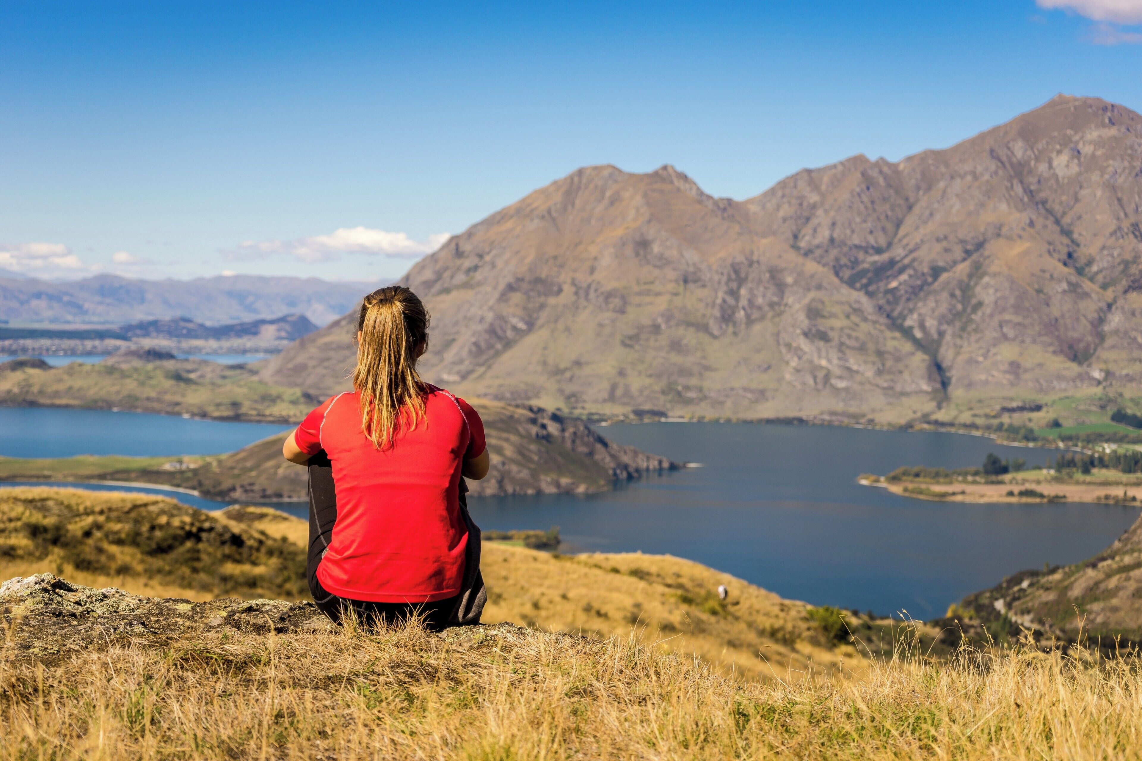 A person in a red shirt sitting on a grassy hill overlooking a lake and mountains.