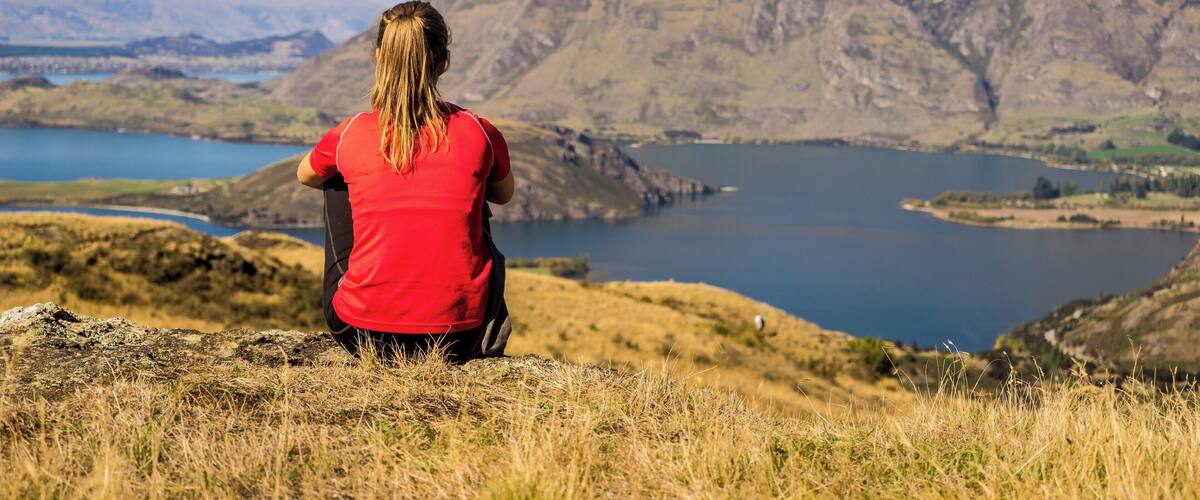 A person in a red shirt sitting on a grassy hill overlooking a lake and mountains.