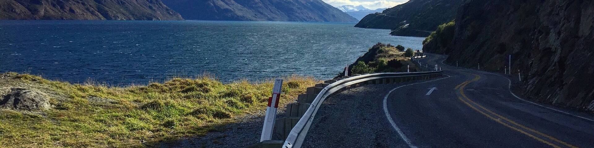 A winding road beside a lake with mountains in the background.
