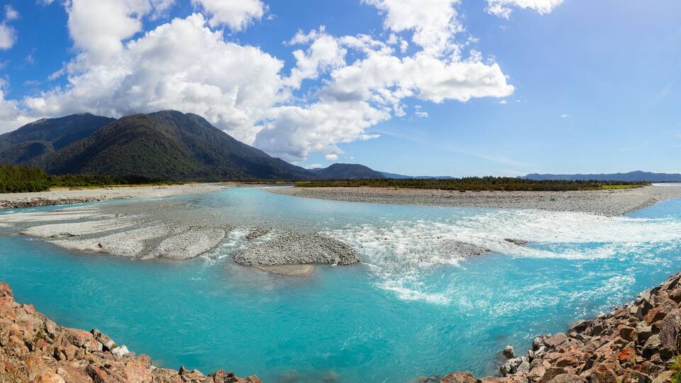 A river with clear turquoise water flowing through a rocky landscape, surrounded by mountains and greenery.