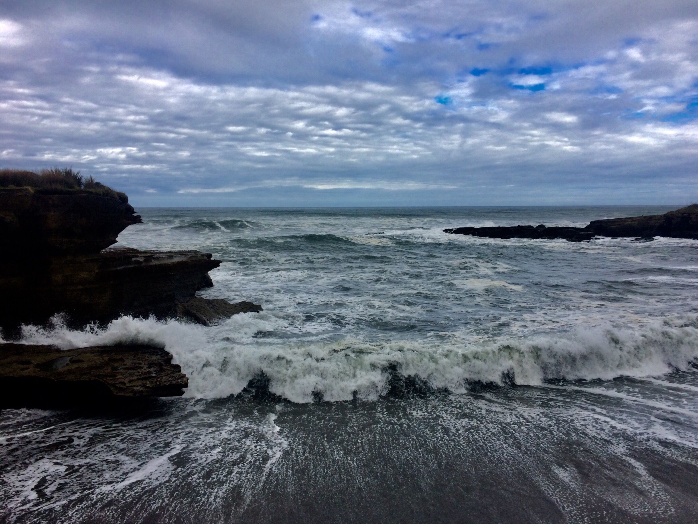 A rocky coastline with waves crashing against the shore under a cloudy sky.
