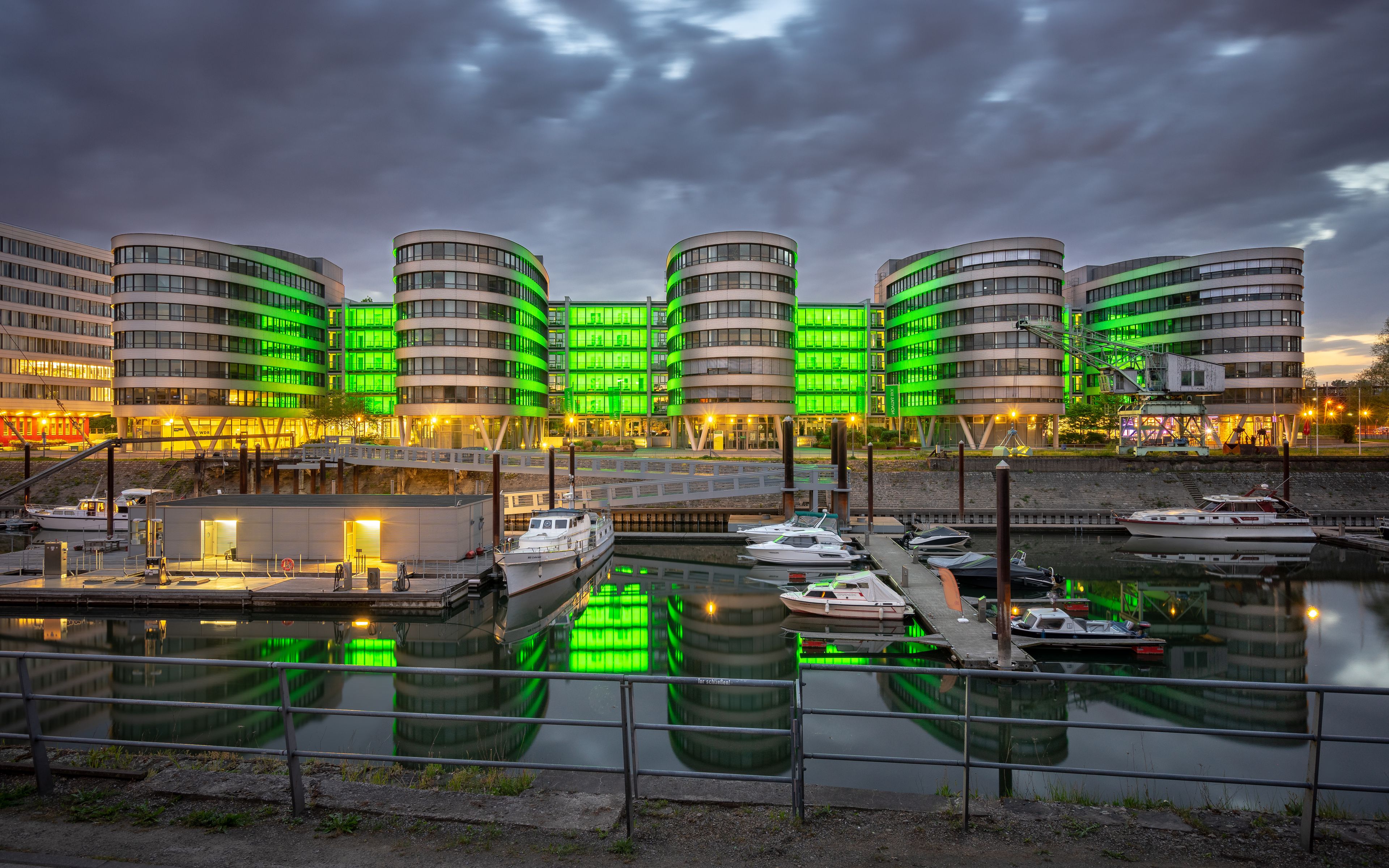 A marina with boats docked, a waterfront promenade, and modern buildings with green lighting.