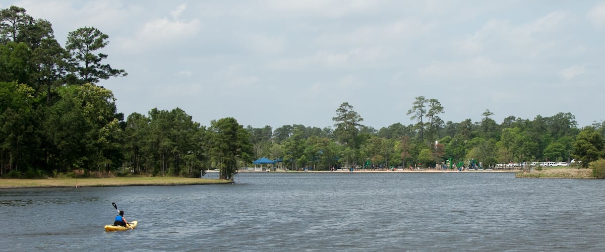 A calm lake with a kayaker, dense trees, and a distant park with structures.