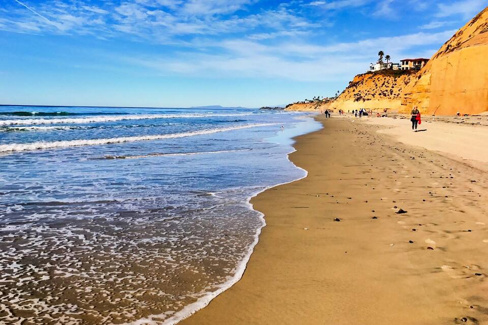 A sandy beach with gentle waves, a clear sky, and a rocky cliff with buildings on top.
