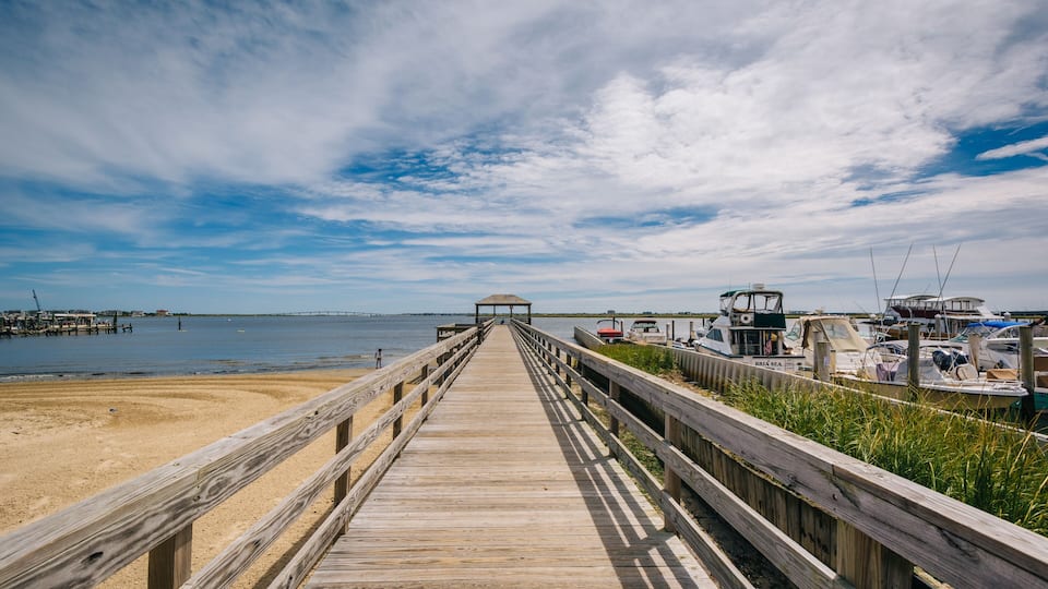 A wooden boardwalk leading to a gazebo on a beach with boats docked nearby.
