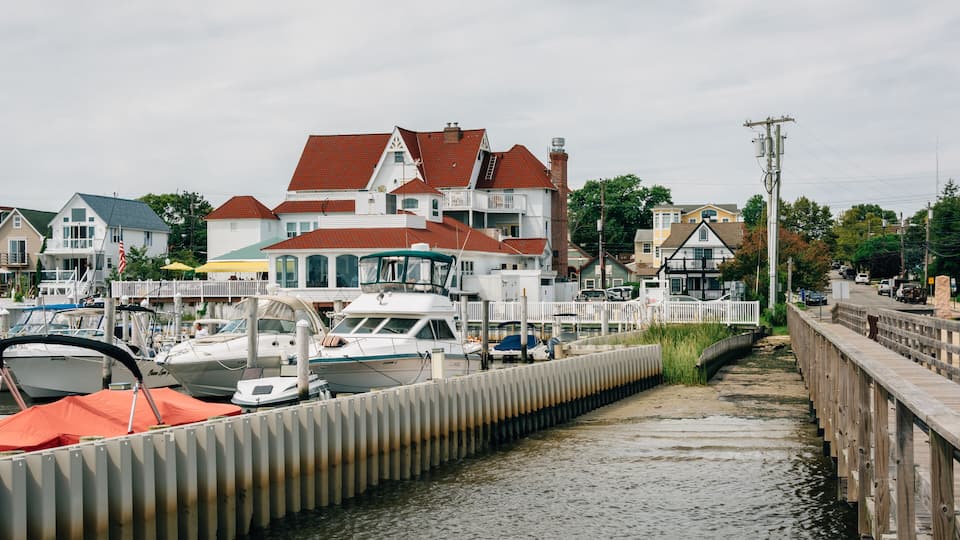 A marina with boats docked, a wooden pier, and houses with red roofs in the background.
