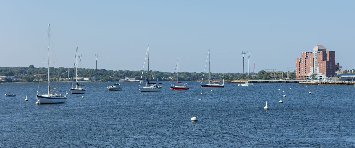 A marina with several sailboats and yachts, a red building, and a distant shoreline with trees and buildings.
