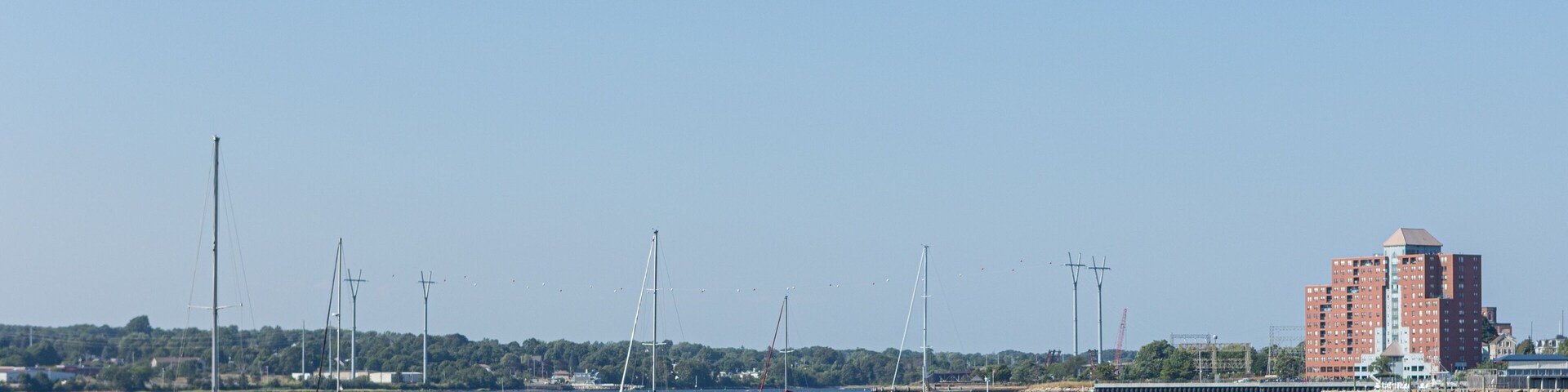 A marina with several sailboats and yachts, a red building, and a distant shoreline with trees and buildings.