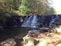 Een waterval die over een stenen muur in een vijver stroomt, omringd door rotsen en bomen.