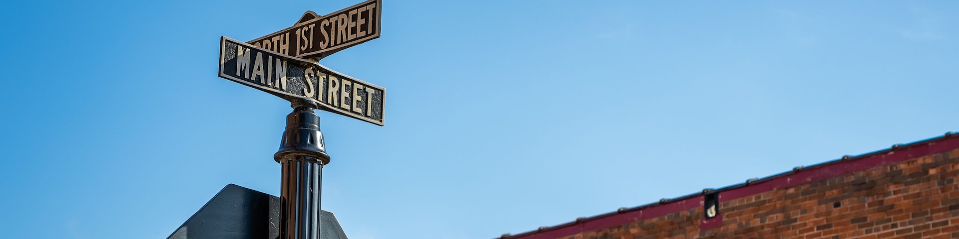 Una señal de calle muestra 'North 1st Street' y 'Main Street', montada en un poste de metal, con un cielo azul despejado y un edificio de ladrillo al fondo.